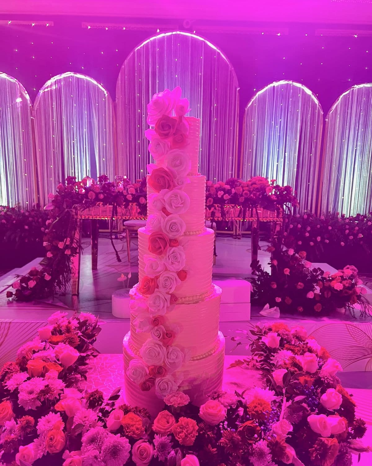Five-tier blush wedding cake lit from behind, flowers running up the back of the tiers